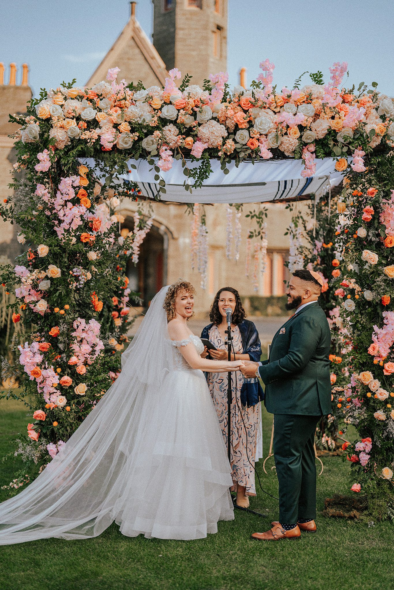 Joyful Jewish wedding ceremony at Whitby Castle, with the bride laughing under a floral chuppah.