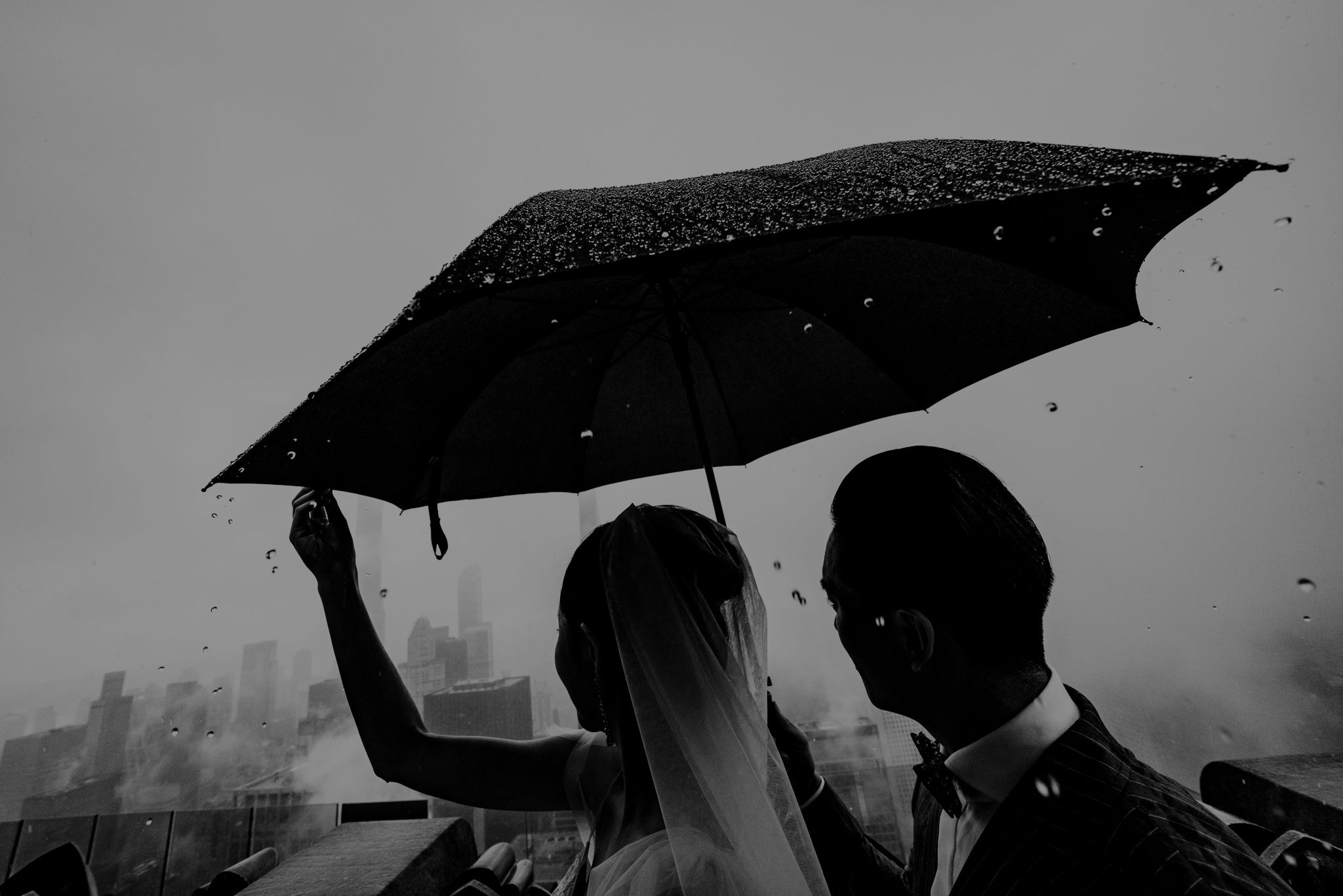Rainy Top of the Rock pre-wedding portrait in black and white, showing the couple sharing an umbrella while overlooking the NYC skyline—moody, cinematic, and intimate.