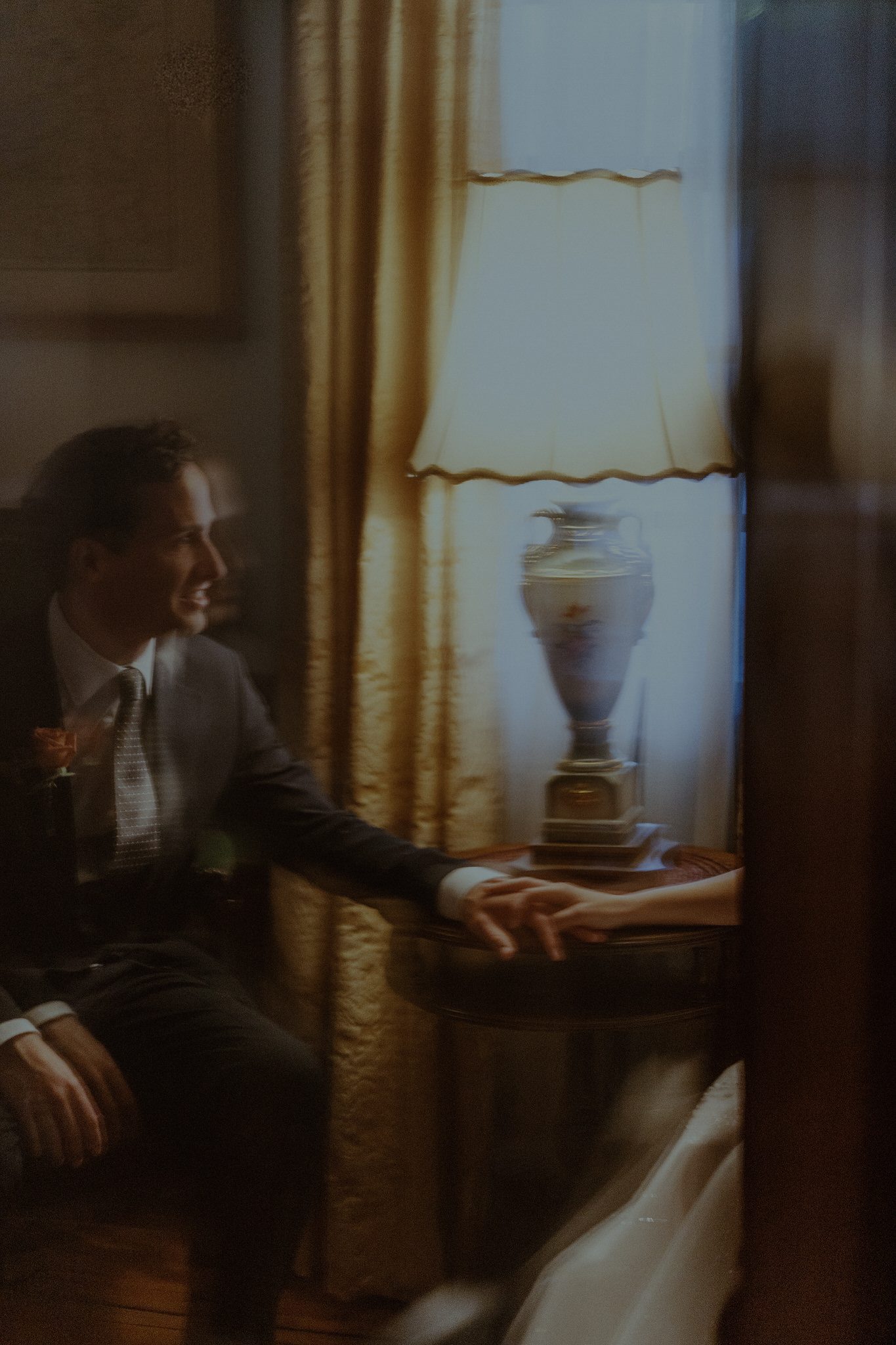 Cinematic wedding portrait of the groom reaching for the bride’s hand beside a warm vintage lamp, photographed through reflections for an intimate, artistic storytelling feel.